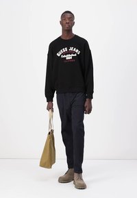 Felpa nera con stampa "GUESS JEANS", abbinata a pantaloni scuri e stivali beige. Tiene una borsa a spalla marrone chiaro. Design semplice e moderno.