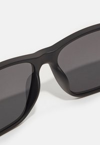 Occhiali da sole neri con un design elegante e angolare, caratterizzati da lenti oscurate e una finitura opaca sulla montatura, che mette in risalto contorni audaci.