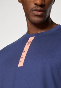 Nike t-paita tummansinisen kankaan, jossa on suuri pystysuuntainen vaaleanpunainen logoprintti edessä. Lyhyet hihat ja pyöreä pääntie.
