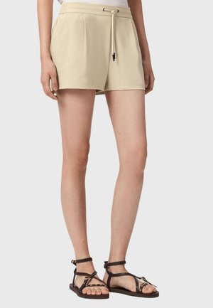 Short beige taille haute avec plis sur le devant et cordon, associé à des sandales noires à bride cheville cloutées, sur des jambes debout sur fond uni.
