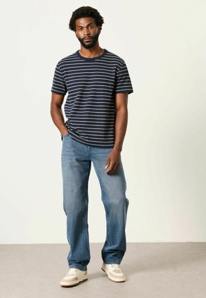 Mann steht mit einer Hand in der Tasche, trägt ein marineblau gestreiftes T-Shirt, blaue Jeans und weiße Turnschuhe vor einfachem Hintergrund.