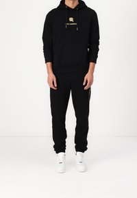 Felpa nera con logo dorato e cordini, abbinata a pantaloni jogger neri con tasca laterale, indossata con sneaker bianche.