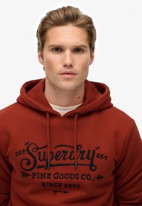 Sudadera roja con capucha ajustable y un estampado gráfico negro en la parte frontal que dice "Superdry Fine Goods Co. Desde 2003." Textura suave.