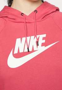 Červená bavlněná mikina s velkým bílým logem Nike, stahovací šňůrkou s černými koncovkami a texturovaným povrchem látky.
