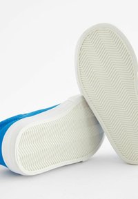 Niebieskie płócienne trampki z teksturowaną białą gumową podeszwą w wzór rybiej ości. Projekt jest niskotopowy z zaokrąglonymi krawędziami.