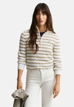 Vrouw met lang bruin haar draagt een beige-wit gestreepte trui met een kwart-ritssluiting en witte broek, houdt een opgevouwen geruite jas in één hand.