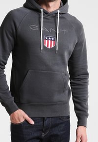 Felpa grigia con cappuccio, dotata di tasca a marsupio, cordini bianchi e logo a scudo con tre stelle in rosso, bianco e blu.