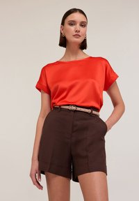 Blouse en satin rouge avec des manches roulées, associée à un short marron avec une taille ceinturée et des poches latérales. Texture lisse et coupe ajustée.