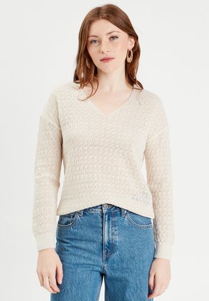 Pull en crème à col en V, en tricot avec un design texturé et ajouré, manches longues, associé à un jean bleu taille haute.
