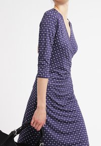 Námořnická modrá šaty s tříčtvrtečními rukávy, které mají diamantový vzor a přiléhavý střih, doplněné černou kabelkou.