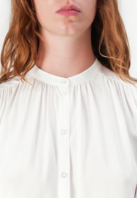 Hvit bluse med høy krage, med myke folder og fem gjennomsiktige knapper foran, laget av lett stoff.