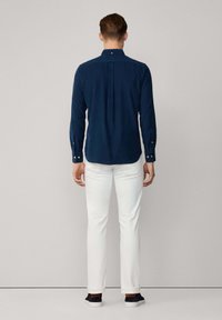 Marineblauw overhemd met lange mouwen en een button-down kraag, met een rugplooi, gecombineerd met witte broek en zwarte sneakers, gezien vanaf de achterkant.