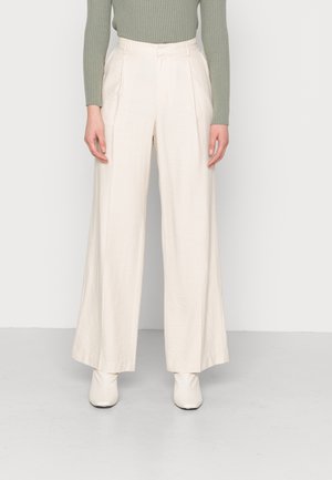 Kobieta ubrana w kremowe, wysokie spodnie o szerokich nogawkach, zielony prążkowany top z długim rękawem oraz białe botki, stojąca na jednolitym tle.