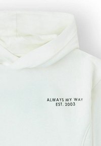 Sudadera blanca con capucha doblada y texto negro en la parte frontal que dice "SIEMPRE A MI MANERA EST. 2003" en la parte superior izquierda del pecho.