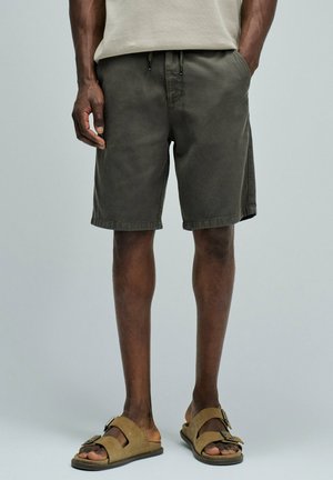 Hombre con pantalones cortos gris oscuro con cordón y sandalias abiertas color tostado con dos tiras de hebilla, de pie frente a un fondo sencillo.