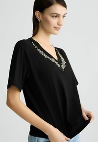 T-shirt nera a V con maniche corte, decorata con pietre decorative lungo il collo, realizzata in tessuto morbido e con una vestibilità rilassata.