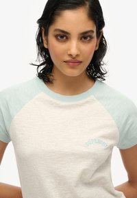 Camiseta de manga corta gris claro con mangas en verde menta y "SUPERDRY" bordado en azul claro. Tejido suave y texturizado.
