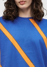 Γυναίκα με καστανά σγουρά μαλλιά που φοράει μπλε πουκάμισο με διαγώνιες πορτοκαλί ρίγες στο στήθος και τον ώμο.