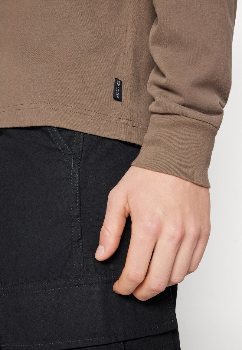 Braune Baumwollbluse mit langen Ärmeln und seitlichem Etikett, getragen über strukturierten schwarzen Hosen mit detaillierter Naht. Sauberer Schnitt und schlichtes Design.