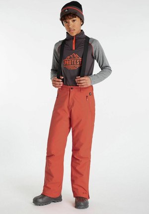 Pantaloni da sci rossi con bretelle nere regolabili, abbinati a una maglietta grigia a maniche lunghe con un logo arancione. Berretto di maglia nero indossato.