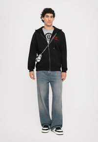 Hanorac negru cu fermoar și broderie roșie, tricou grafic gri dedesubt, blugi largi de culoare albastru deschis și adidași alb-negru.