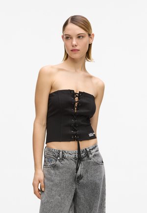 Modèle féminin portant un haut corset noir à lacets sans bretelles avec logo et un jean gris délavé taille haute, debout devant un fond uni.