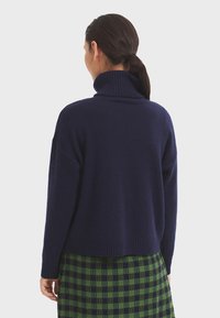 Marineblauer Rollkragenpullover mit lockerer Passform, gerippten Bündchen und Saum. Getragen über einem grün-schwarz karierten Rock. Weiche Textur.
