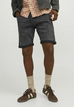 Shorts in denim scuro con orlo risvoltato, abbinati a una camicia a fantasia e sneakers marroni con strisce crema, completati da calze beige.