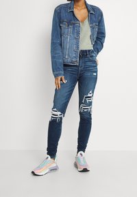 Jeansjacka, grå V-ringad topp, slitna blå jeans med fransade knän och pastell sneakers med flerfärgade accenter och svarta sulor.