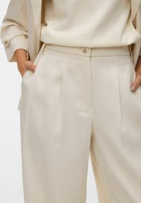 Beżowe, dopasowane spodnie o gładkiej fakturze, z plisami, bocznymi kieszeniami i zapięciem na guzik. Noszone z jasnym topem.