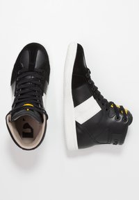 Černé vysoké tenisky vyrobené z kůže a semiše s bílými akcenty, kulatá špička, texturovaná gumová podrážka a kontrastní žlutý detail na jazyku.