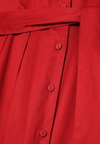 Vestido de tela roja con diseño plisado, cierres laterales con botones y un cinturón a juego. La textura parece suave con un ligero brillo.