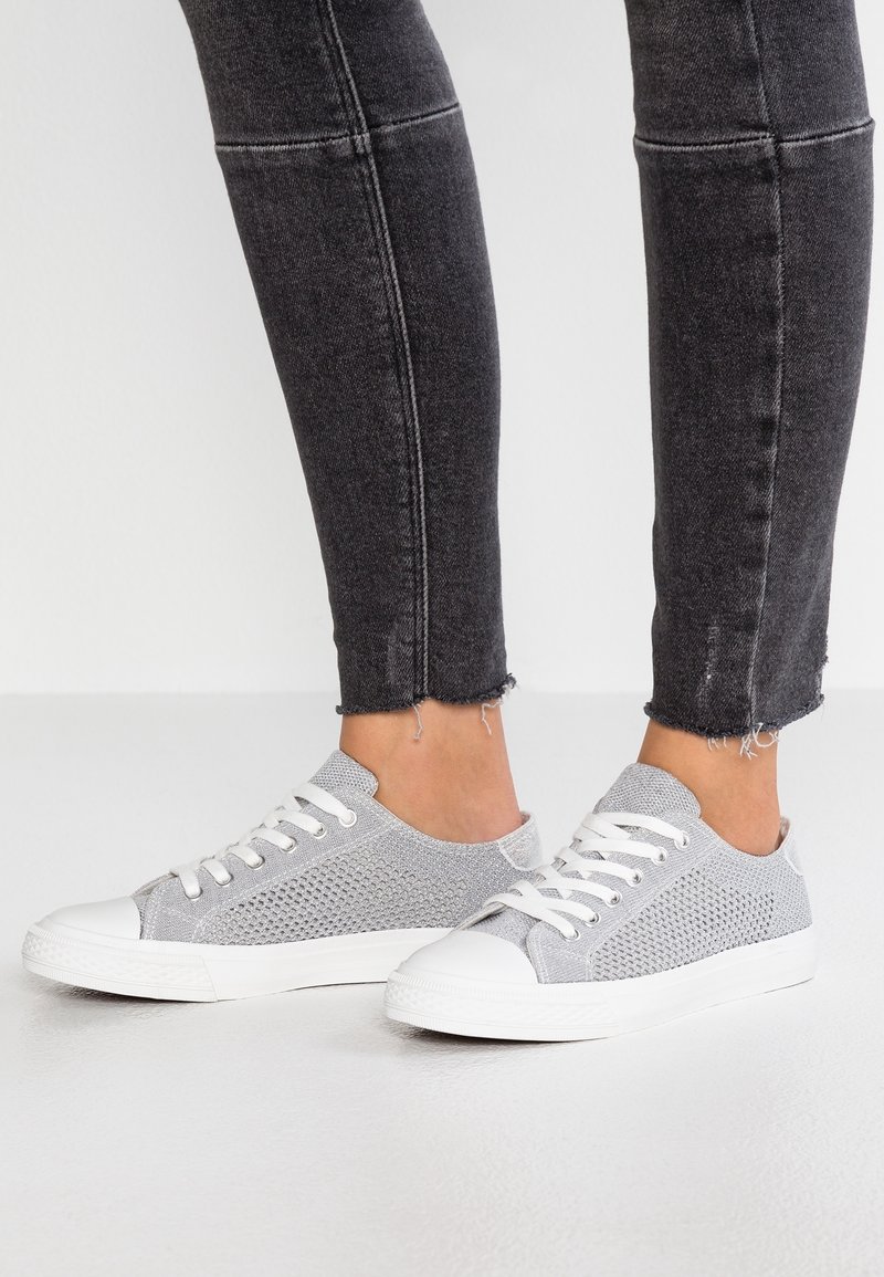 Harmaat verkkokengät, joissa on valkoiset kumiset varvaskupit ja nauhat. Ominaisuuksiin kuuluu teksturoitu päällinen ja matala profiili. Yhdistetty mustien, röpelöisten farkkujen kanssa.