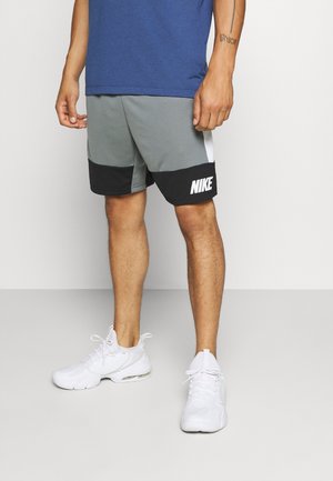 Uomo che indossa pantaloni corti Nike grigi e neri, sneakers bianche e una maglietta blu, in piedi contro uno sfondo bianco semplice.