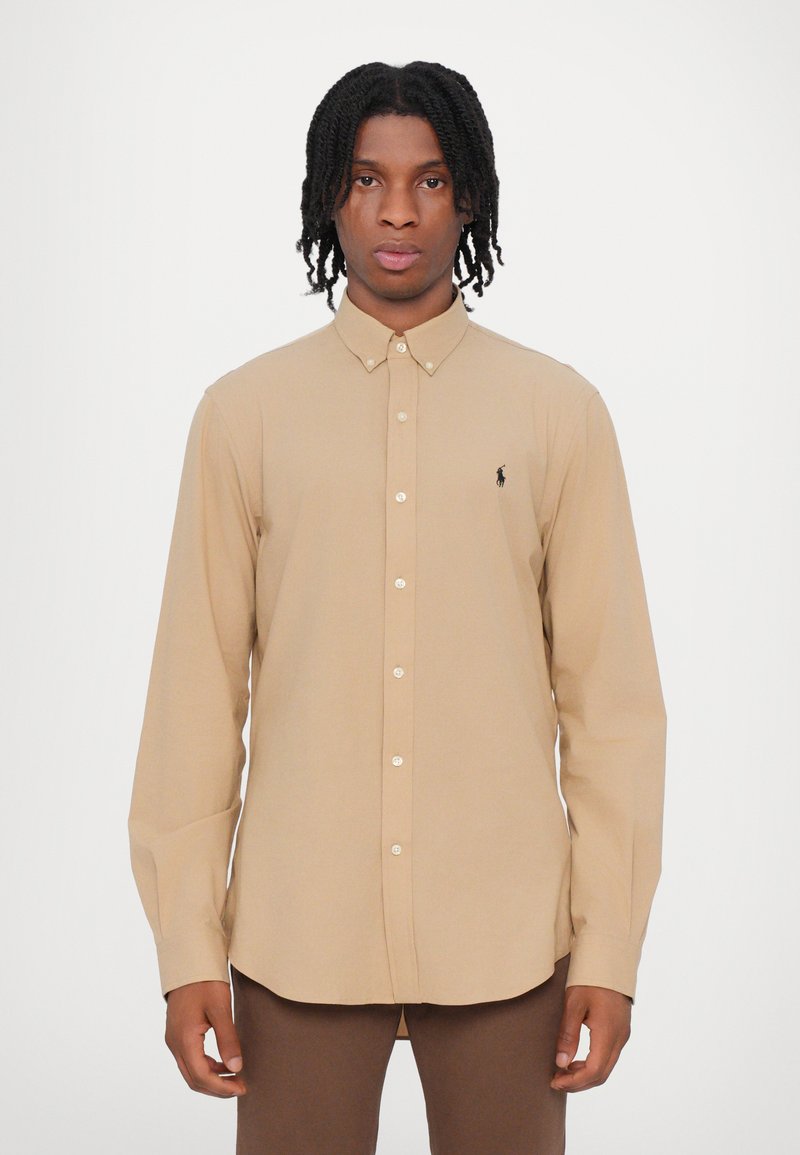 Camisa bege com colarinho, mangas longas e um pequeno logótipo preto no peito. Tecido suave e corte clássico.