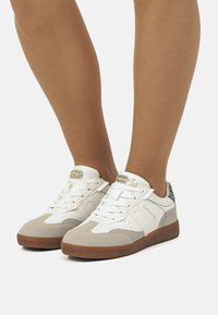 Zapatillas blancas y gris claro con acentos de ante, diseño de cordones, cuello acolchado y suela de goma texturizada. Con una etiqueta de logo metálico.