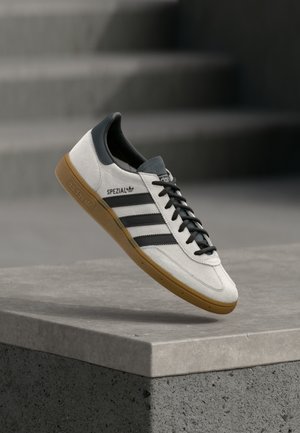Baskets blanches Adidas Spezial avec des bandes noires et une semelle beige posées au-dessus d'un rebord en béton gris.