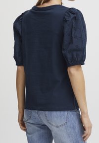 Haut bleu marine à manches bouffantes en tissu texturé, orné de broderies florales. Le dos présente un design simple et lisse.