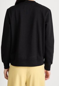 Svart sweatshirt med rund halsringning, ribbad manschetter och nederkant. Slät textur, enkel design och avslappnad passform. Bärs med gula byxor.