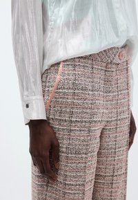 Pants in pink and black tweed with a textured pattern, featuring side pockets and a light pink accent. Accompanied by a sheer, light-colored blouse.