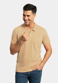 Camisa polo beige de manga corta, con abertura y logo bordado en el pecho. Combinada con jeans azul oscuro y una pulsera de plata.