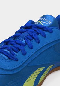 Κοντινή λήψη ενός μπλε αθλητικού παπουτσιού Reebok με φωτεινά μπλε κορδόνια και λογότυπο σε κίτρινο-πράσινο χρώμα στο πλάι, με σόλα από καουτσούκ χρώματος καραμέλας.