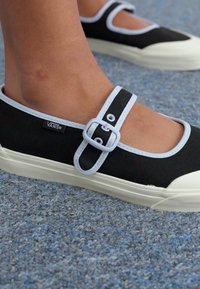 Sapatos de tela pretos com um acabamento azul claro e uma fita de fivela, apresentando um design clássico de perfil baixo e uma sola de borracha texturizada.