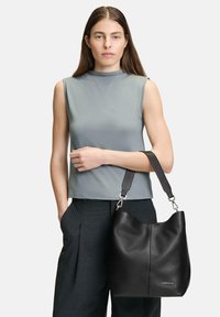 Schwarze Leder-Hobo-Tasche mit gewölbtem Oberteil, silbernem Metallzubehör und breitem Schultergurt, gehalten von einem Model in einem grauen ärmellosen Oberteil.