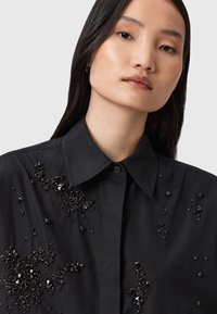 Giovane donna con lunghi capelli scuri che indossa una camicia nera con colletto decorata con intricate applicazioni di perline nere.