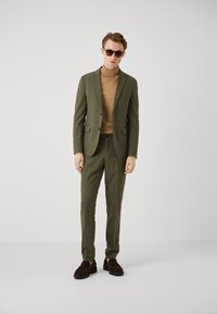 Olive green suit with a single-breasted jacket, matching trousers, and a beige turtleneck. Brown suede shoes and sunglasses complete the look.
