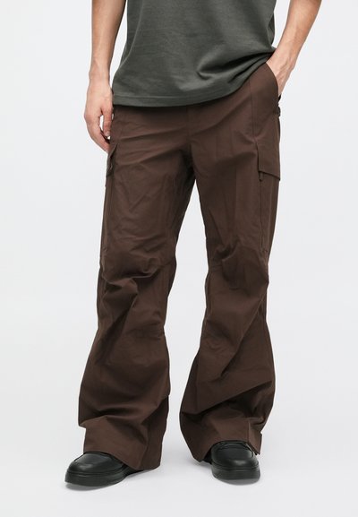 Rjave cargo hlače z ohlapnim krojem, narejene iz lahkega materiala, z stranskimi žepi in diskretno sijočo površino, v kombinaciji s črnimi čevlji.