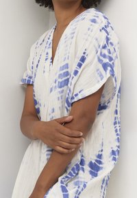 Vit textilskjorta med blå batikmönster, lös passform, V-hals, korta ärmar, texturerad tyg. Modellens armar korsade framför.