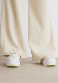 Sneakers bianchi con una suola spessa e piattiforme e dettagli verdi, abbinati a pantaloni larghi beige chiaro con una texture a coste.