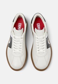 Sneaker bianche in pelle con dettagli neri sui lati, suole marroni, lacci bianchi e solettes rosse contrassegnate con la scritta "CAMPER" in bianco.